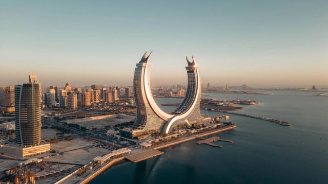 Luftaufnahme des markanten Raffles Doha mit seinen zwei sichelförmigen Türmen direkt an der Uferpromenade, umgeben von modernen Hochhäusern und dem ruhigen Wasser des Arabischen Golfs bei Sonnenuntergang.