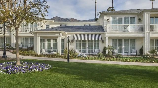 Elegante weiße Gebäude des Rosewood Miramar Beach Resorts mit Balkonen, Veranden und gestreiften Markisen, umgeben von gepflegtem Rasen, Blumenbeeten und Palmen vor einer bergigen Kulisse.