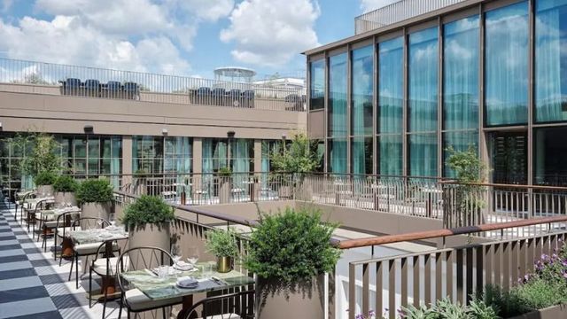 Moderne Dachterrasse des Rosewood Vienna mit eleganten Sitzbereichen, bepflanzten Trennelementen und großflächigen Glasfassaden, unter blauem Himmel mit vereinzelten Wolken – urbanes Flair in stilvollem Ambiente.