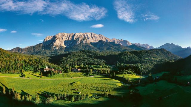 Panoramaaufnahme des luxuriösen Schloss Elmau, eingebettet in eine weitläufige grüne Alpenlandschaft mit Wäldern und Wiesen, vor der eindrucksvollen Kulisse des Wettersteingebirges in Bayern.