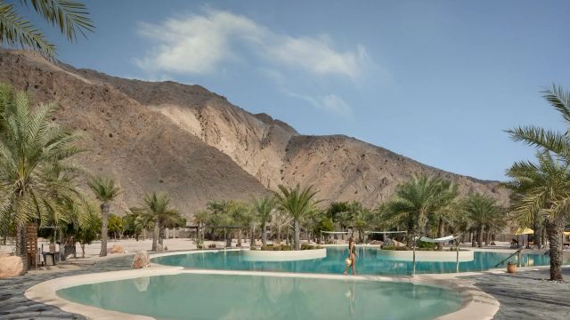 Blick auf den palmengesäumten Pool des Six Senses Zighy Bay Resorts im Oman, umgeben von einer majestätischen Felskulisse und Wüstenlandschaft, mit einer Person, die entspannt am Wasser entlanggeht.