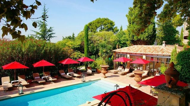 Sonniger Außenbereich eines eleganten Hotels mit rechteckigem Pool, umgeben von bequemen Liegen mit roten Sonnenschirmen, mediterranen Pflanzen und klassischer Architektur.