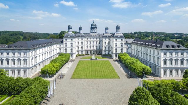  Prächtiges Barockschloss in symmetrischer Architektur mit zentralem Ehrenhof, gepflegten Rasenflächen und umliegenden Baumalleen – das Grandhotel Schloss Bensberg bei strahlend blauem Himmel in erhöhter Lage über dem Bergischen Land.