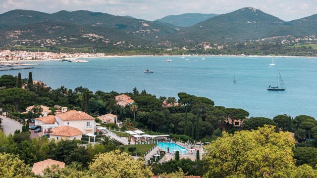 Blick auf die luxuriöse Althoff Villa Belrose an der französischen Riviera bei Saint-Tropez, umgeben von mediterraner Vegetation, mit Poolterrasse im Vordergrund und traumhaftem Blick auf die azurblaue Bucht und bewaldete Hügel im Hintergrund.