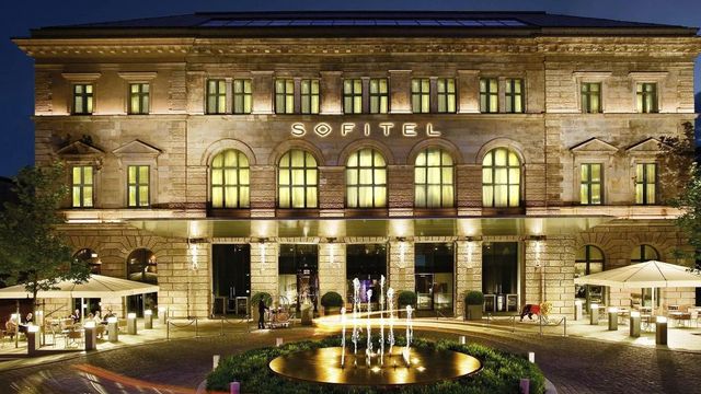 Beleuchtete Fassade des Sofitel Munich Bayerpost bei Nacht mit Springbrunnen.