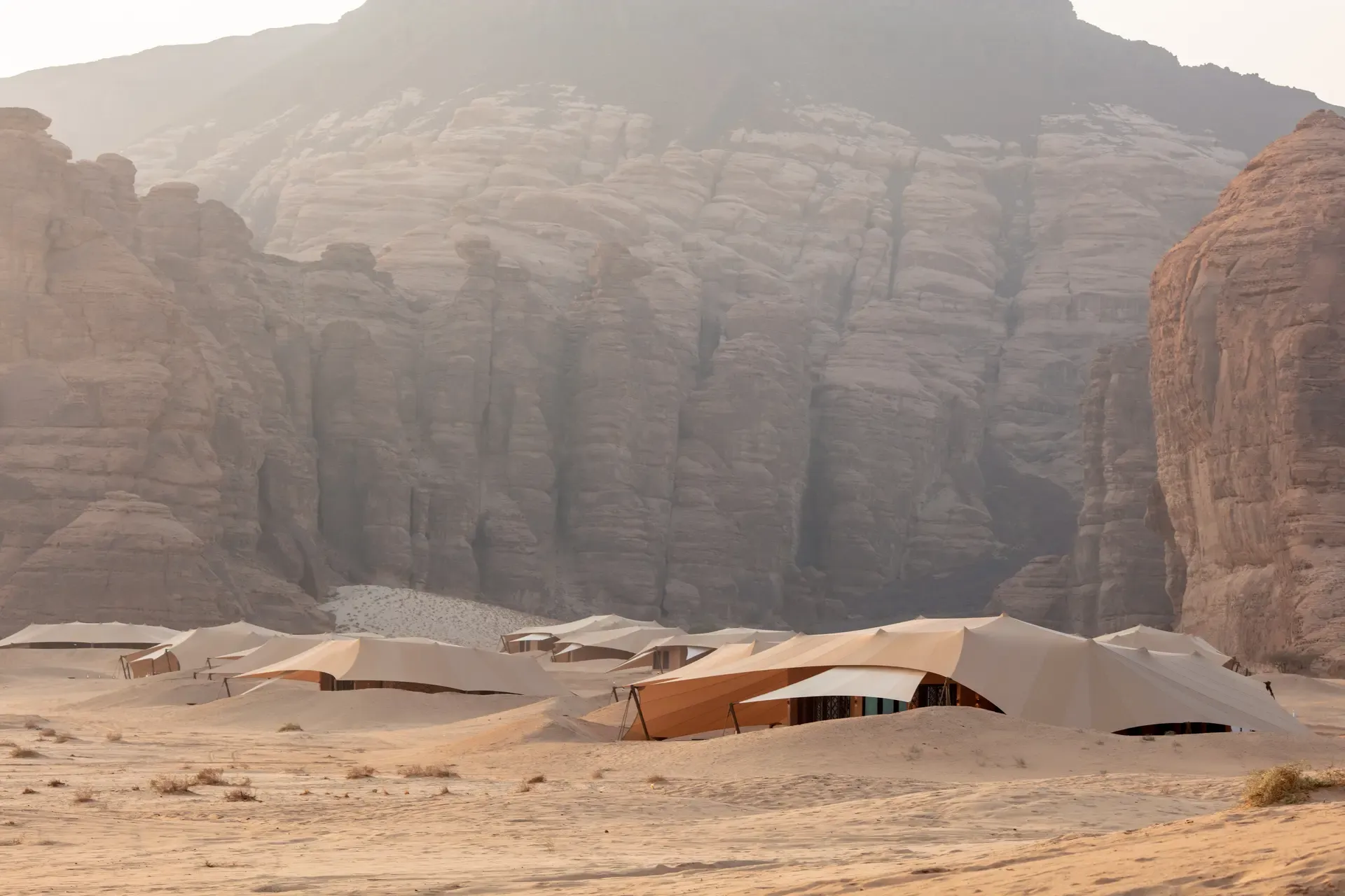 Luxuriöse Wüstenzelte des Banyan Tree AlUla Resorts eingebettet in die sandige, von mächtigen Sandsteinfelsen umgebene Landschaft von AlUla, Saudi-Arabien – in warmem Morgenlicht, das die dramatische Kulisse weich durchleuchtet.