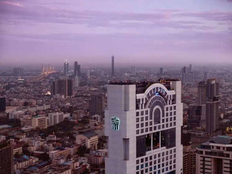 Das Banyan Tree Bangkok ragt als markanter Wolkenkratzer über die Skyline der Stadt, mit seiner ikonischen Architektur und der beliebten Rooftop-Bar Vertigo, die bei Sonnenuntergang beleuchtet ist.