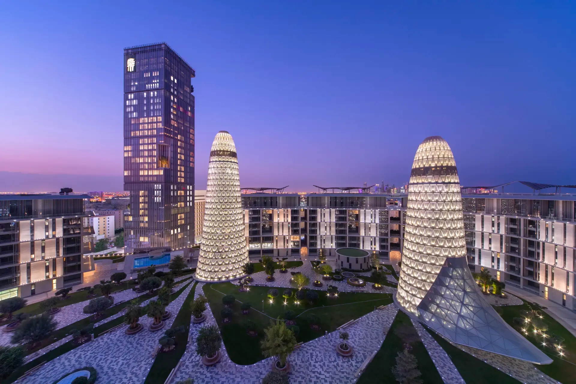 Abendaufnahme des Banyan Tree Doha Resorts im futuristischen Doha Oasis-Komplex, mit beleuchteten, kegelförmigen Glastürmen, modernem Hochhaus und elegantem Innenhof mit begrünten Wegen und luxuriöser Architektur unter violett-blauem Himmel.