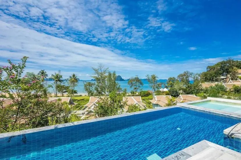 Infinity-Pool mit Blick auf tropische Vegetation, Strand und das türkisfarbene Meer im Banyan Tree Krabi. Im Hintergrund erheben sich imposante Kalksteinfelsen unter einem strahlend blauen Himmel.
