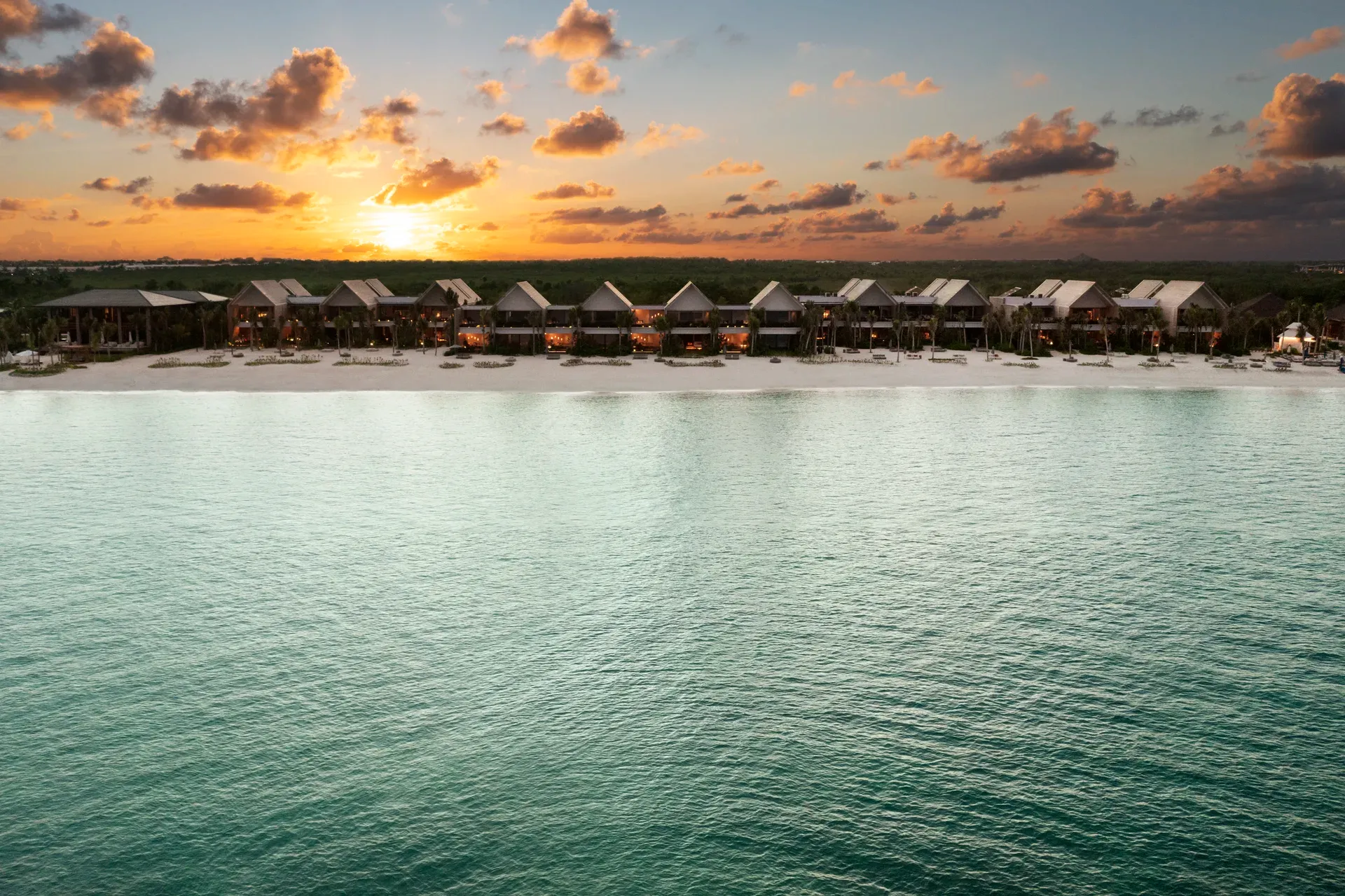 Luxuriöse Strandvillen des Banyan Tree Mayakoba Resorts in Mexiko bei Sonnenuntergang, mit ruhigem türkisfarbenem Meer im Vordergrund und dramatisch gefärbtem Himmel im Hintergrund.