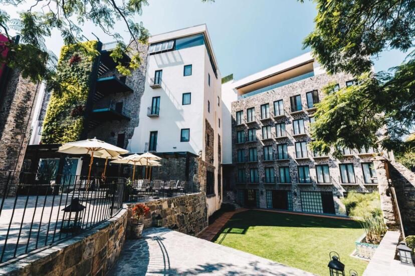 Innenhof des Banyan Tree Puebla mit stilvoller Steinfassade, moderner Architektur, Sonnenschirmen auf der Terrasse und gepflegtem Garten – umgeben von historischem Charme und urbanem Flair.