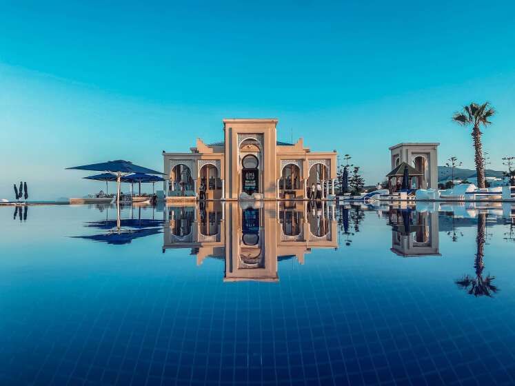 Majestätisches Gebäude im maurischen Stil im Banyan Tree Tamouda Bay, gespiegelt in einem ruhigen Infinity-Pool, umgeben von Palmen und luxuriöser Resort-Atmosphäre bei strahlend blauem Himmel.