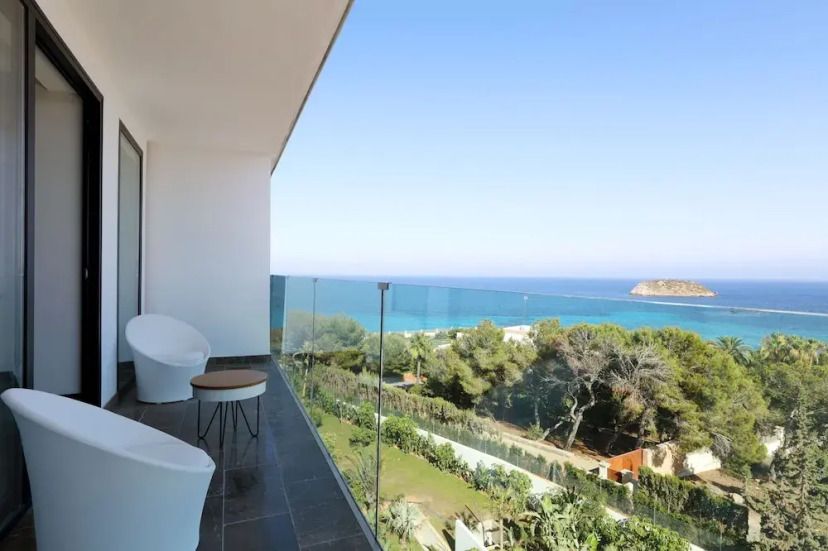 Moderner Balkon mit Glasgeländer im Bless Hotel Ibiza, mit Blick auf das tiefblaue Meer, eine kleine vorgelagerte Insel und grüne Landschaft.