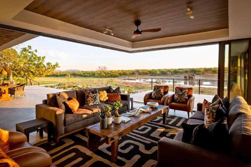 Offene Lounge der Jamala Madikwe Royal Safari Lodge mit eleganten Sofas, Holztisch, afrikanischem Dekor und atemberaubendem Blick auf ein Wasserloch mit Elefanten – luxuriöses Safari-Ambiente in freier Natur.
