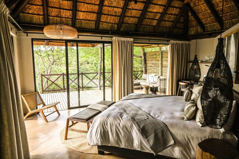 Gemütliche Suite der Laluka Safari Lodge mit Reetdach, stilvollem Interieur, großem Bett, natürlichen Materialien und Panoramafenstern, die auf eine private Holzterrasse mit Blick ins Grüne führen.