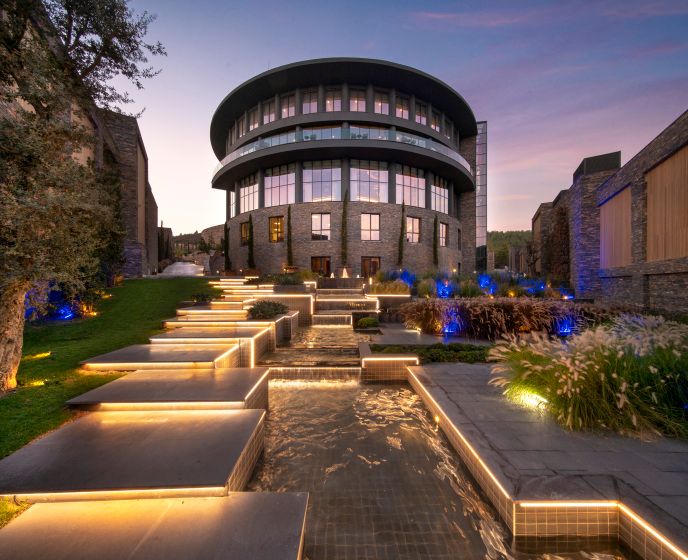 Modernes Rundgebäude des Lujo Bodrum Hotels bei Sonnenuntergang, mit terrassenförmigen Wasserbecken und beleuchteten Stufen, umgeben von gepflegtem Garten und eleganter Architektur aus Stein und Glas.