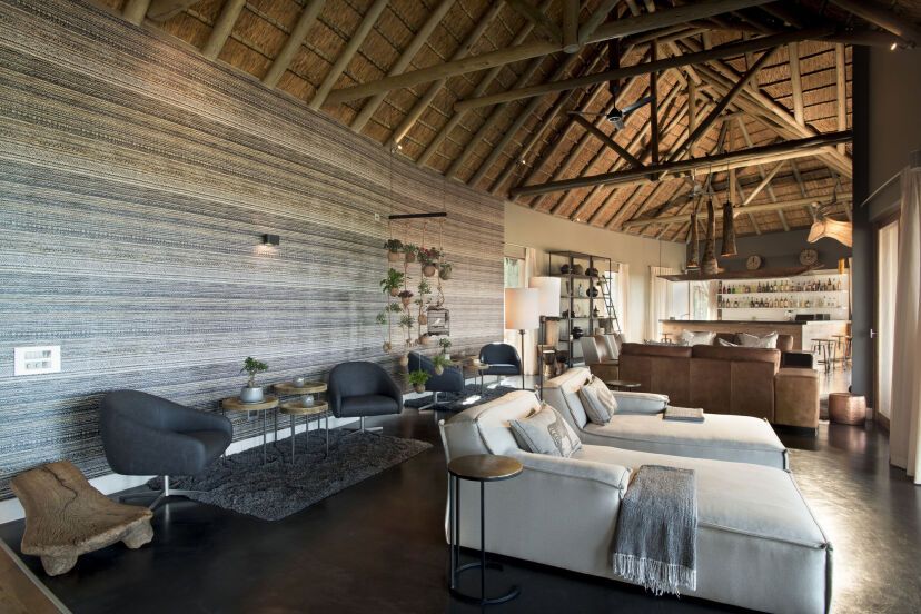 Stilvoller Lounge- und Barbereich der Mhondoro Safari Lodge & Villa mit modernen Liegen, gemütlichen Sesseln, offener Reetdachkonstruktion und elegantem Interieur im afrikanischen Design.