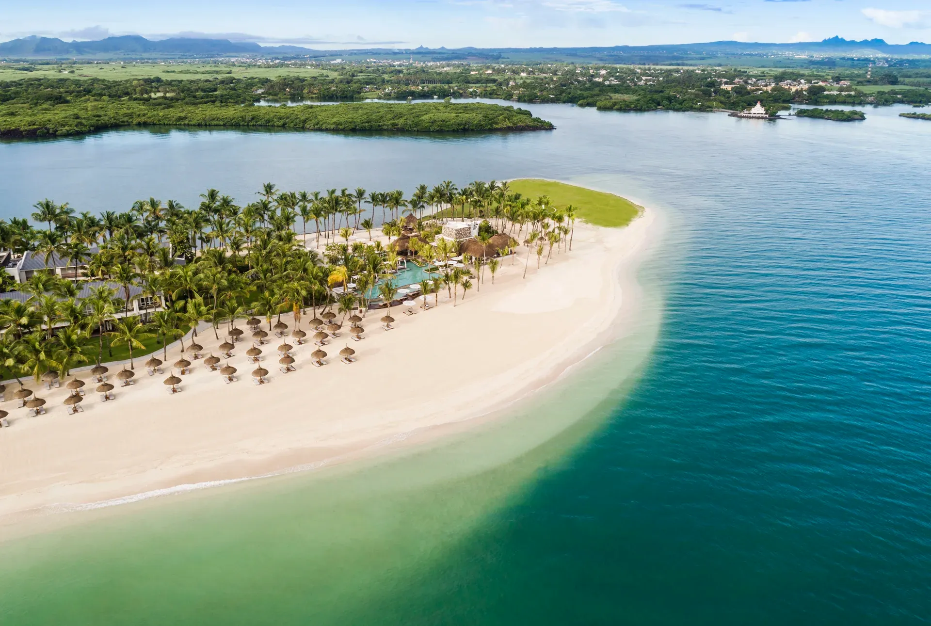  Luftaufnahme eines tropischen Luxusresorts mit strohgedeckten Sonnenschirmen, Palmen und einem weißen Sandstrand auf einer schmalen Landzunge in Mauritius, umgeben vom türkisblauen Indischen Ozean und einer üppig grünen Landschaft im Hintergrund.