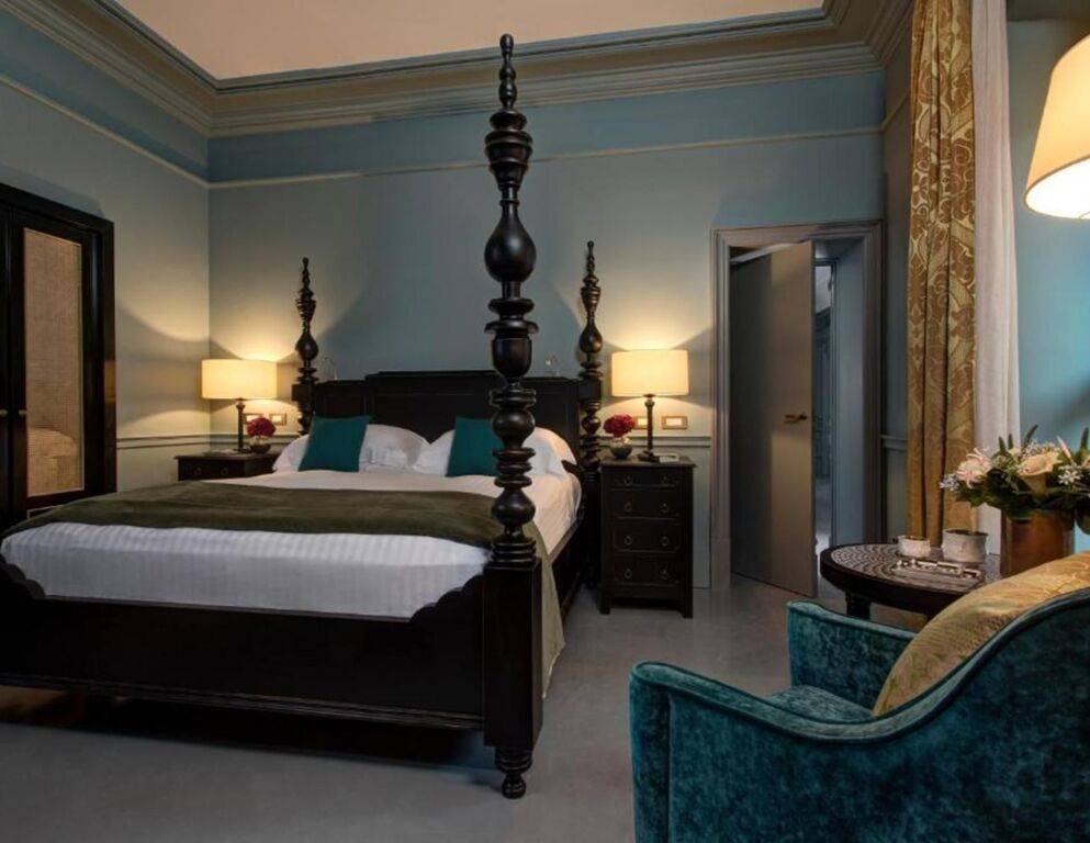 Alt-Text: Stilvolles Schlafzimmer in der Rocco Forte House mit Himmelbett aus dunklem Holz, eleganter Beleuchtung, samtigem Sessel und klassisch dekoriertem Interieur.