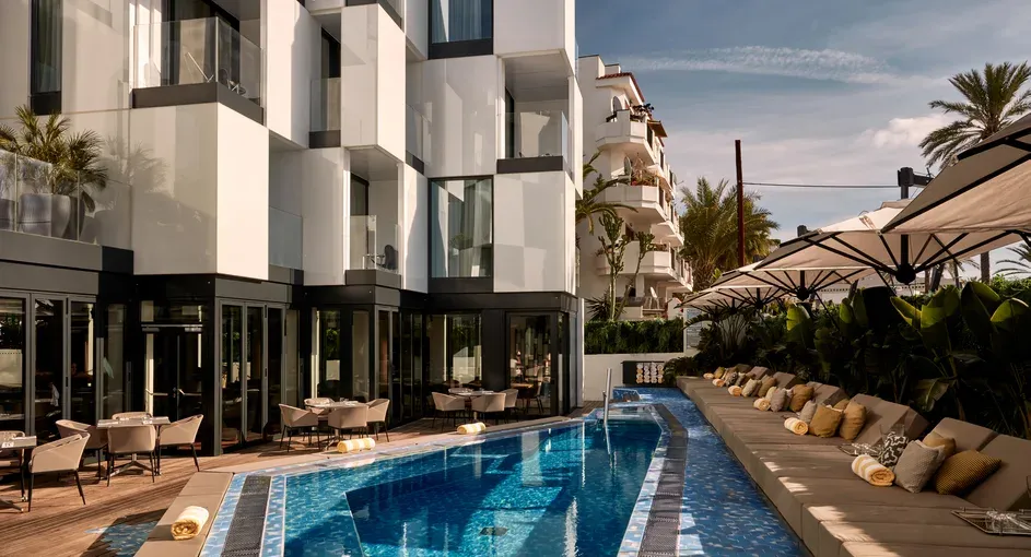Moderner Außenbereich des Sir Joan Hotels in Ibiza mit kantiger Architektur, einem schmalen Pool und stilvollen Sonnenliegen unter Sonnenschirmen, umgeben von Palmen und eleganter Atmosphäre.