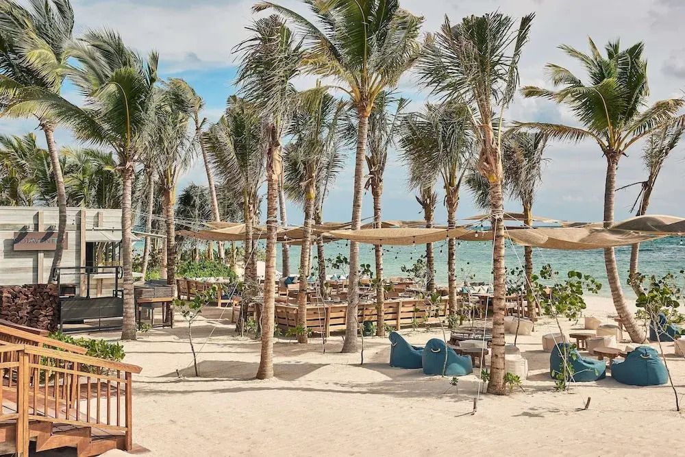 Strandbereich mit Palmen und Loungemöbeln am Meer, entspannte Atmosphäre.