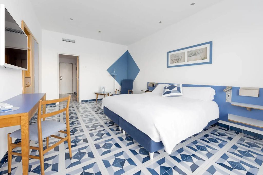 Helles Hotelzimmer mit blau-weißem geometrischen Bodenmuster und Doppelbett