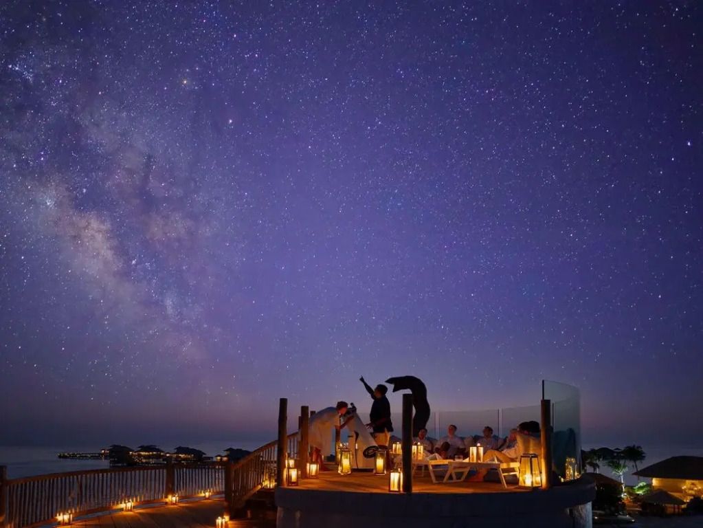 Sternenhimmel über einer beleuchteten Terrasse mit Teleskop und Gästen