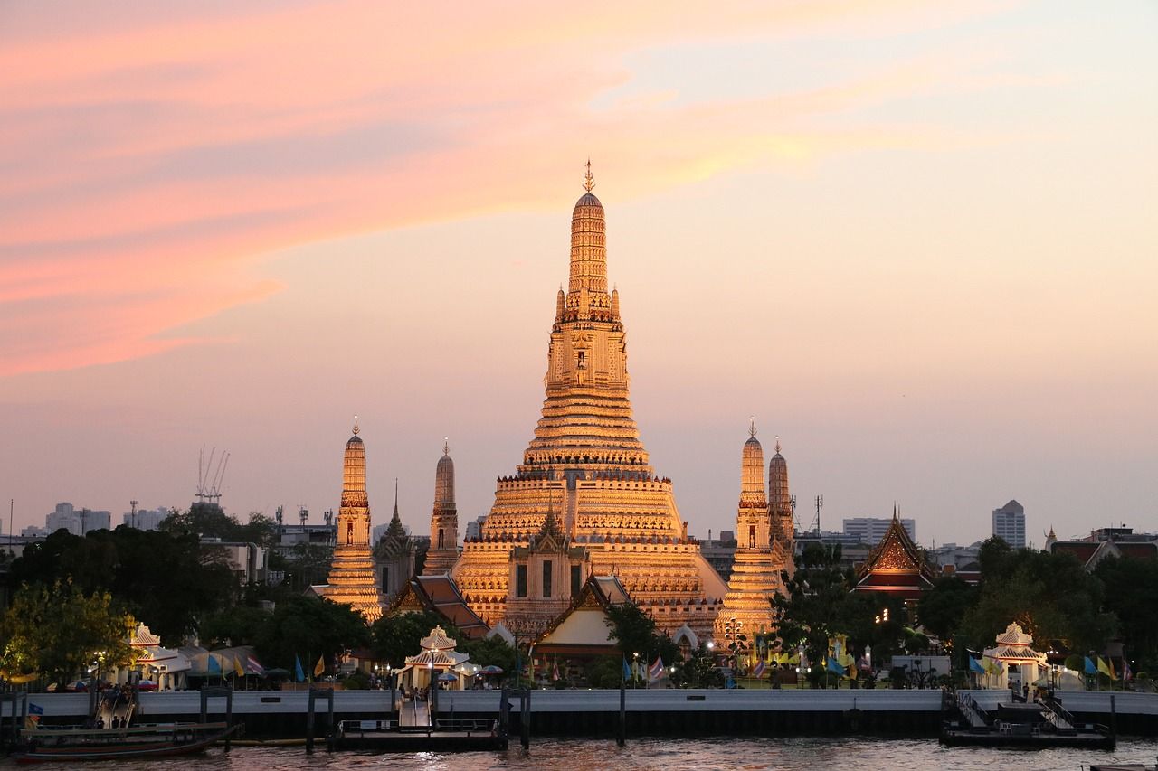 Der Wat Arun erstrahlt im warmen Licht des Sonnenuntergangs, seine filigranen Türme spiegeln sich sanft im ruhigen Fluss und schaffen eine magische, friedvolle Atmosphäre.